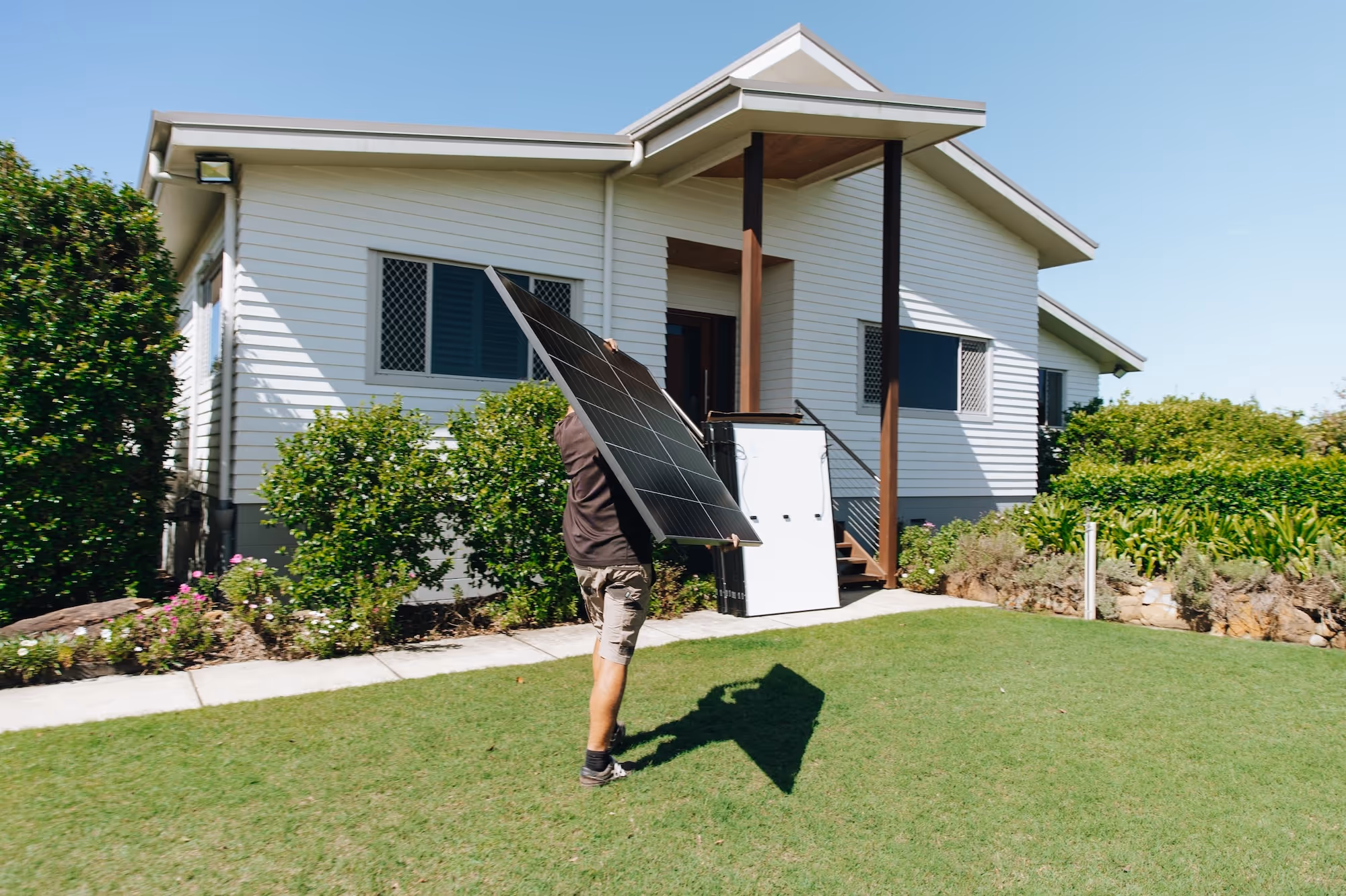 An LMS Electrical installer carrying a solar panel across the lawn towards a Queensland home.